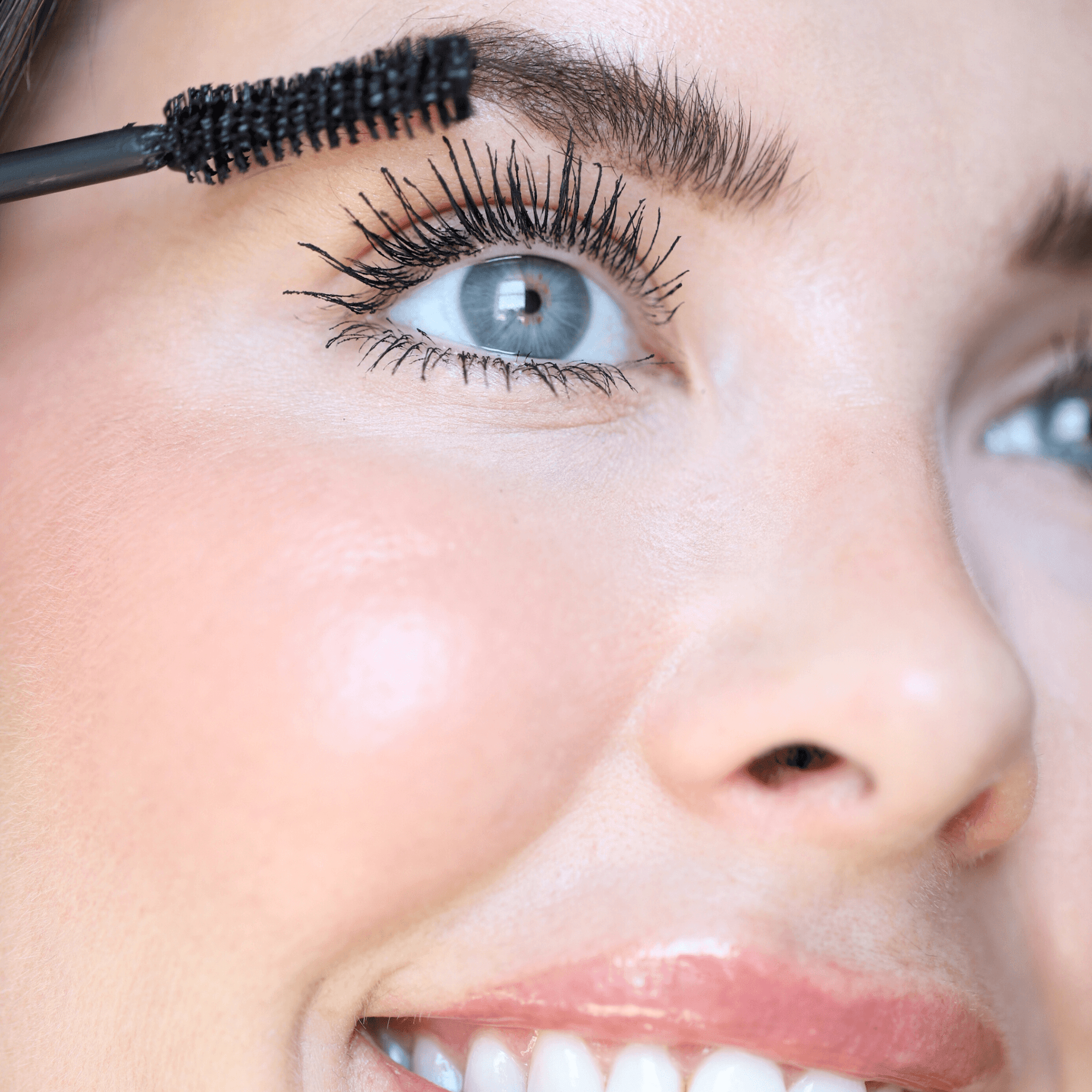 Close-up of a smiling person applying Eye Defy Zero Gravity Mascara to upper eyelashes, showcasing the mascara wand's hourglass shape and the product's clump-free, volumizing effect.