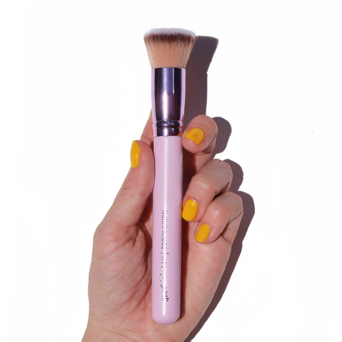 shiny purple flat-top foundation kabuki in hand with yellow fingernails - complexion brush- half caked
